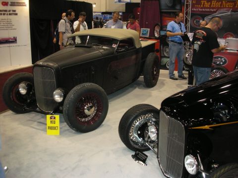 1998 Ford Crown Victoria - Two early Ford-style hot rods, a flat-black roadster with wire wheels and a glossy black coupe, displayed at an indoor show.