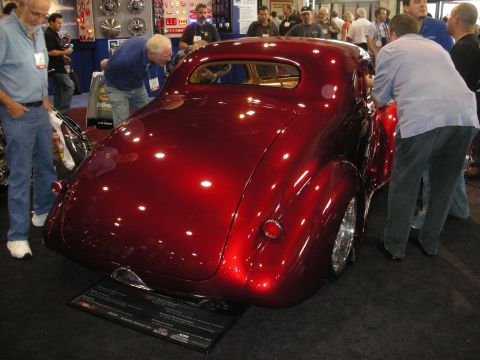 1998 Ford Crown Victoria - rear of a deep candy-red custom coupe with chrome wheels draws onlookers around a placard on an indoor show floor.