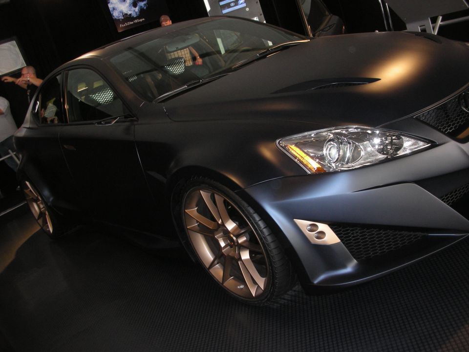 Front-quarter view of matte-black Lexus coupe with bronze wheels under show lighting
