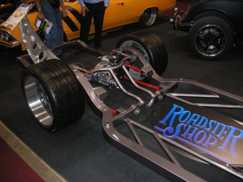 1998 Ford Crown Victoria - bare tubular chassis with independent suspension and wide tires sits on display next to a Roadster Shop logo panel at an indoor show.