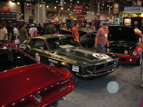 1998 Ford Crown Victoria - black YearOne Track Car Mustang fastback wearing number 67 sits among other show cars on a crowded convention floor.