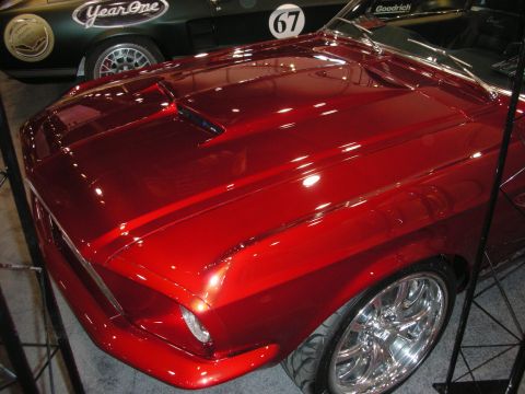 1998 Ford Crown Victoria - deep candy-red fastback hood with raised scoops gleams under show lights, with a number-67 race car visible behind it.