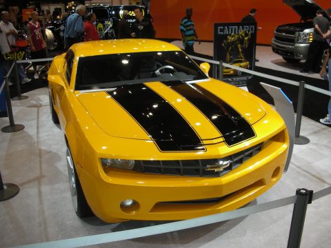 1998 Ford Crown Victoria - yellow Chevrolet Camaro with twin black racing stripes sits behind stanchions at an auto show beside a Camaro display banner.
