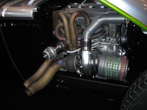 1998 Ford Crown Victoria - polished supercharged engine with intricate twisting exhaust headers and a finned blower mounted in the bay of a black hot rod chassis.