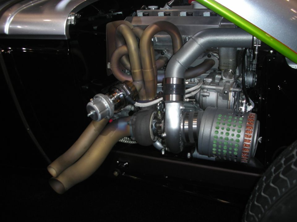Close-up of a chrome-and-silver supercharged engine with twisting exhaust headers in a hot rod chassis