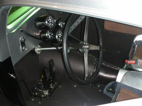 1998 Ford Crown Victoria - spartan interior of a custom hot rod showing a large black steering wheel, a cluster of round black-faced gauges, and exposed metal panels.