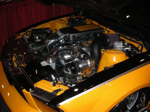 1998 Ford Crown Victoria - chrome belt-driven supercharged engine with a red cone air filter sits in the bay of a yellow car at an indoor show.