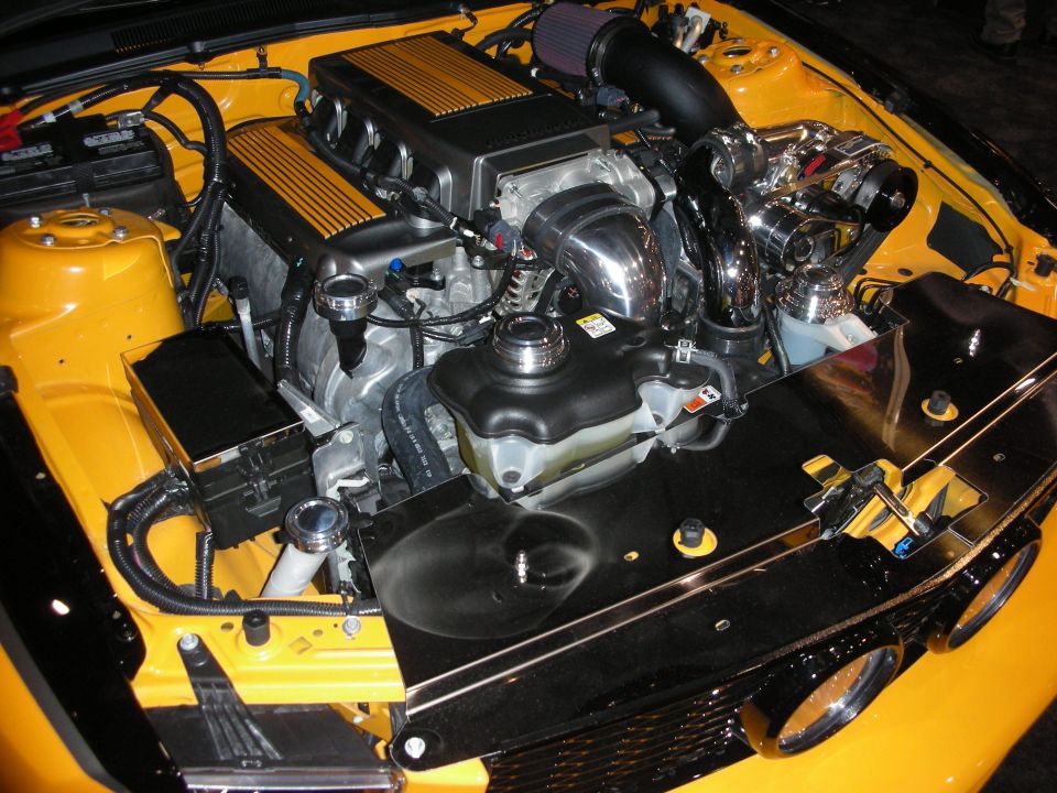 Engine bay of a yellow car with finned cam covers, chrome supercharger and detailed plumbing