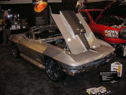 1998 Ford Crown Victoria - silver mid-1960s Chevrolet Corvette convertible displayed with its hood open on a show stand beside a red truck.