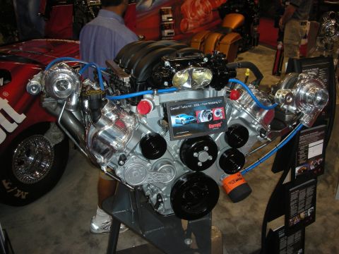 1998 Ford Crown Victoria - twin-turbo crate V8 on a display stand with blue plumbing, chrome turbos and Garrett signage at an indoor show.