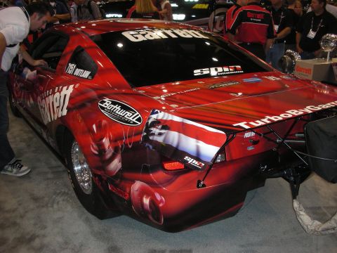 1998 Ford Crown Victoria - Rear three-quarter of a red drag car wrapped in Garrett turbo livery with airbrushed graphics and a rear parachute mount at a show.
