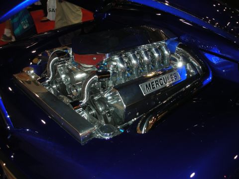 1998 Ford Crown Victoria - heavily chromed blown V8 engine set into the smoothed blue hood opening of a custom car, with a Mercules plate on the intake.