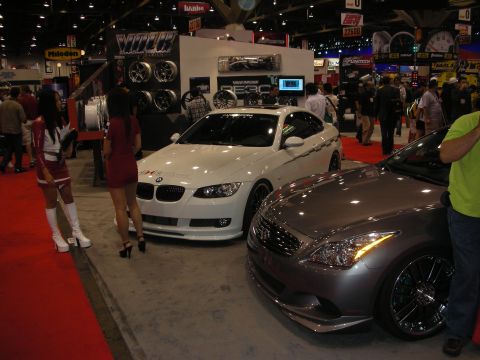 1998 Ford Crown Victoria - white BMW coupe and a silver Infiniti coupe displayed near a wheel booth on red carpet at a crowded indoor trade show.