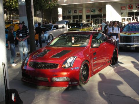 1998 Ford Crown Victoria - Candy-red widebody Infiniti G35 coupe with a vented hood and black wheels, displayed outdoors among a crowd of onlookers.