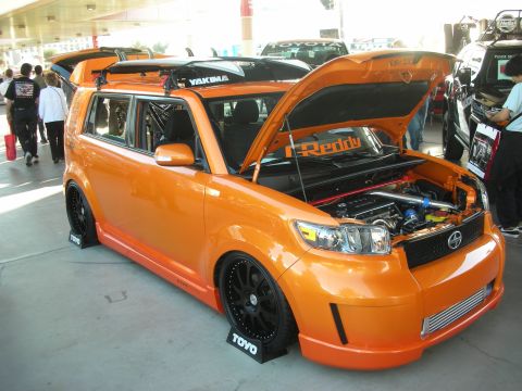 1998 Ford Crown Victoria - Lowered orange Scion xB box wagon with open hood revealing a turbo setup and roof rack, displayed at an outdoor car show.