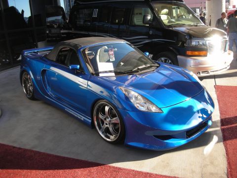 1998 Ford Crown Victoria - Blue custom Toyota MR2 Spyder roadster with widebody bodywork and chrome wheels, parked on red carpet beside a van.