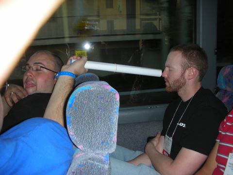 1998 Ford Crown Victoria - Two men sit on a bus at night; one in blue holds a white rolled tube toward a bearded man in a black tee wearing a lanyard.