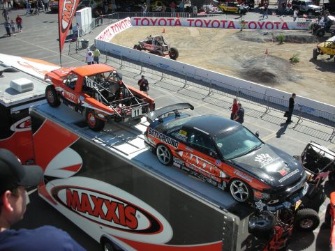 1998 Ford Crown Victoria - Maxxis-branded display trailer holding an orange off-road race truck and a black liveried drift car at an outdoor show.