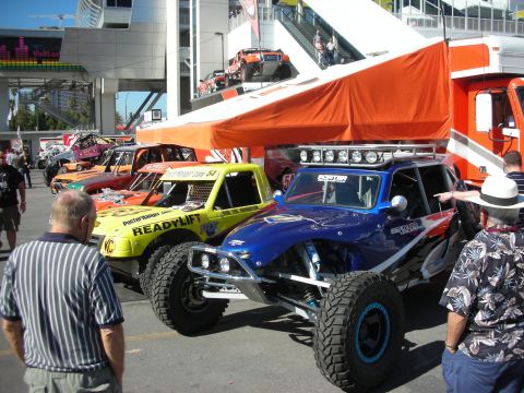 1998 Ford Crown Victoria - Row of colorful off-road desert race trucks and buggies parked under an orange canopy as spectators walk past.