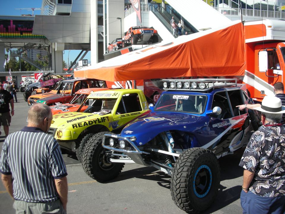 Row of off-road desert race trucks under an orange canopy at an outdoor show