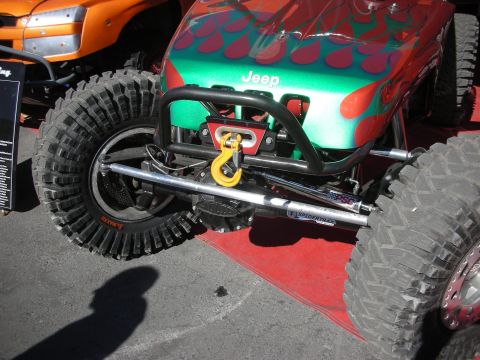 1998 Ford Crown Victoria - Close-up of a green-and-red flamed Jeep rock crawler's front suspension, tubular bumper, winch hook and chunky mud tires.
