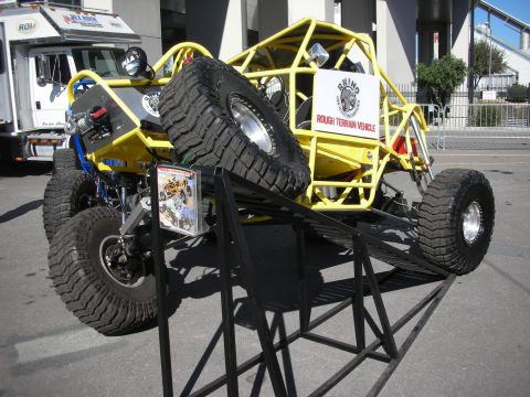 1998 Ford Crown Victoria - Yellow tube-frame rough-terrain buggy with large off-road tires, displayed tilted on a metal stand at an outdoor show.