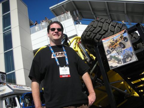 1998 Ford Crown Victoria - Smiling man in a black tee and sunglasses, wearing a SEMA buyer lanyard, posing beside a tilted yellow off-road buggy.