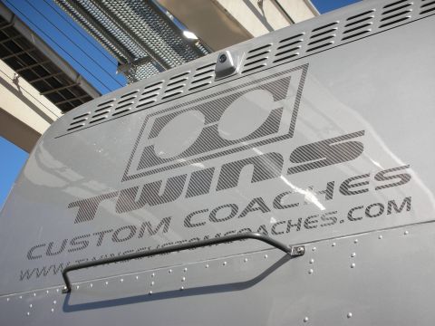 1998 Ford Crown Victoria - silver coach side panel displays the 'DC Twins Custom Coaches' logo and website, photographed under an overpass against blue sky.