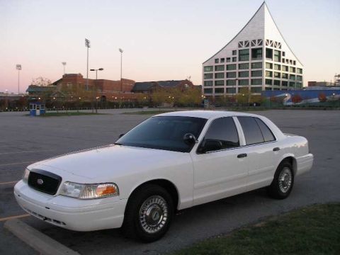 2001 Ford Crown Victoria - A white 2001 Ford Crown Victoria sedan is parked in an empty lot with a modern building in the background.