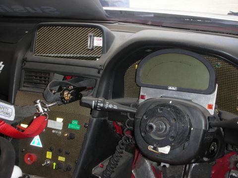 1998 Ford Crown Victoria - stripped race-car cockpit showing a carbon-fiber dash, MoTeC digital display, steering hub, and safety harness webbing.