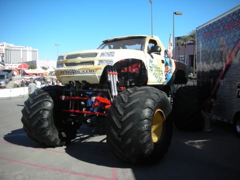 1998 Ford Crown Victoria - white-bodied monster truck on massive flotation tires sits in a parking lot with city buildings in the background.