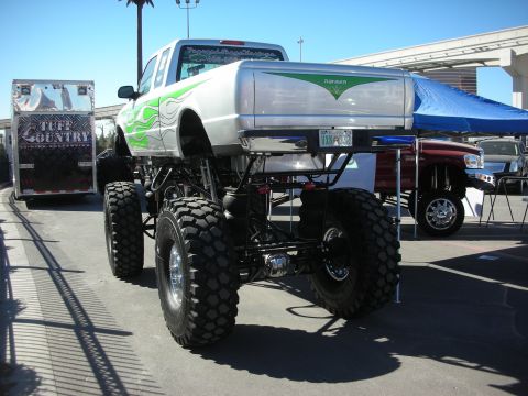 1998 Ford Crown Victoria - silver pickup-bodied monster truck with green flames and tall lift sits on giant tires beside a Tuff Country trailer.