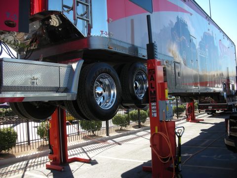 1998 Ford Crown Victoria - long enclosed car-hauler trailer raised on red two-post lifts, exposing its underside and polished dual wheels.