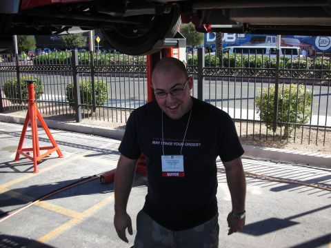 1998 Ford Crown Victoria - smiling bald man in glasses and a black t-shirt with a 'Buyer' event badge stands beneath a vehicle on a lift.
