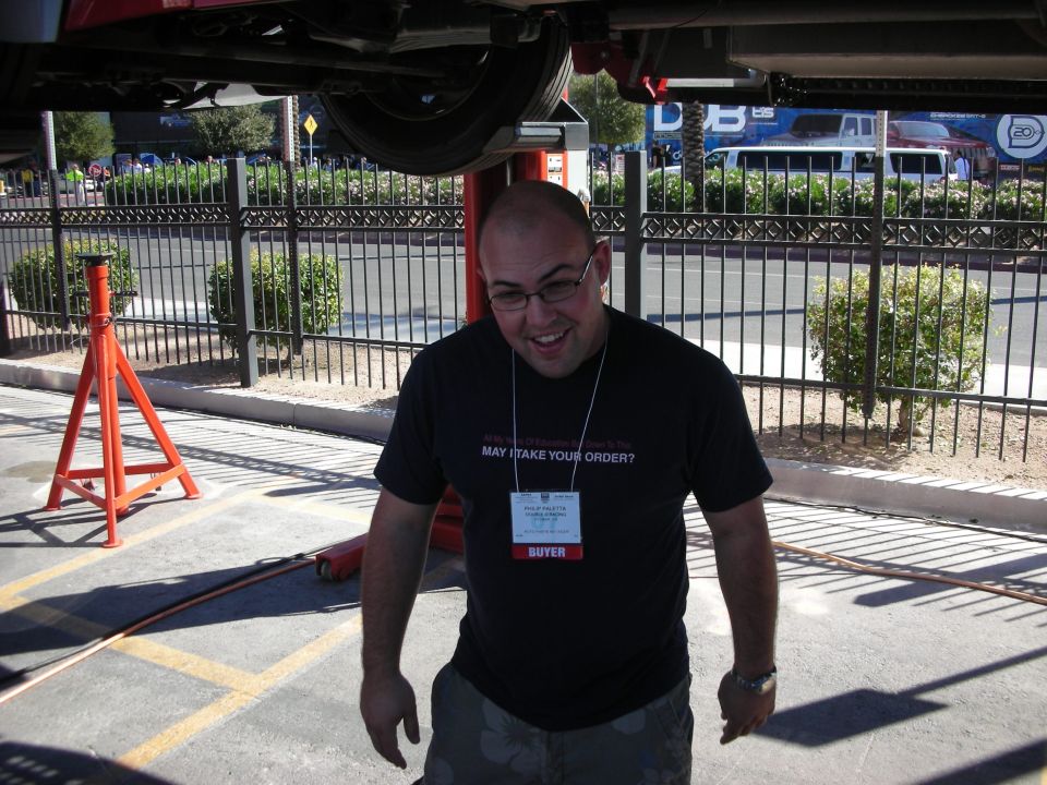 Smiling bald man in a black t-shirt with event badge standing under a raised vehicle