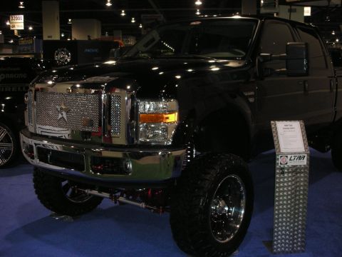 1998 Ford Crown Victoria - lifted black Ford Super Duty pickup with a chrome diamond-plate grille, big mud tires and chrome wheels displayed at an indoor show.