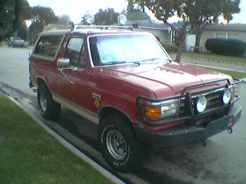 1989 Ford Bronco - This red Ford Bronco is parked on a residential street with a bull bar and auxiliary lights on the front.