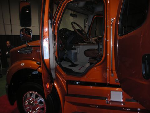 1998 Ford Crown Victoria - orange custom medium-duty truck with its door open, revealing the seats and steering wheel, parked on a show floor.