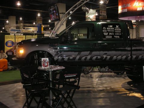 1998 Ford Crown Victoria - dark green lifted pickup with 'Geldermann' rocker graphics and sponsor decals displayed with director's chairs at an indoor expo booth.