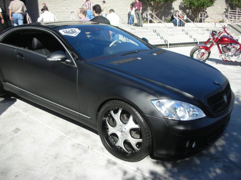 1998 Ford Crown Victoria - matte black luxury coupe on chrome wheels parked outdoors on pavement, with a custom chopper motorcycle and crowd in the background.