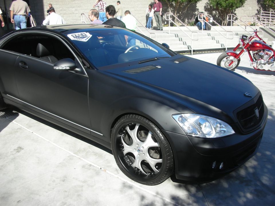 Matte black luxury coupe with chrome wheels parked outdoors near a chopper