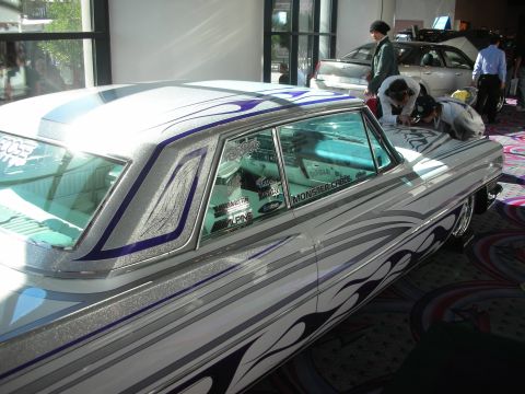 1998 Ford Crown Victoria - silver custom car with purple accents and pinstriped flames, with people leaning over it while detailing at an indoor venue.