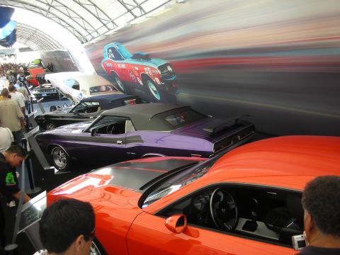 1998 Ford Crown Victoria - row of classic muscle cars, including orange and purple coupes, displayed under a curved exhibition roof with motion-blur backdrop.