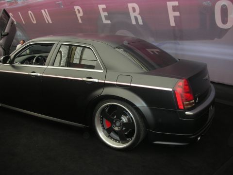1998 Ford Crown Victoria - Matte-black custom Chrysler 300 sedan, rear three-quarter view on large multi-spoke wheels with red brake calipers at an indoor display.