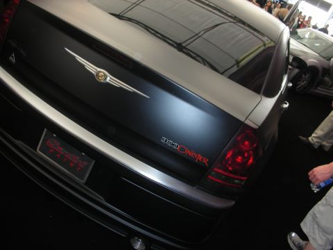 1998 Ford Crown Victoria - Trunk of a matte-black Chrysler 300 showing the winged Chrysler emblem and a red '300 Sinister' graphic above the taillight.