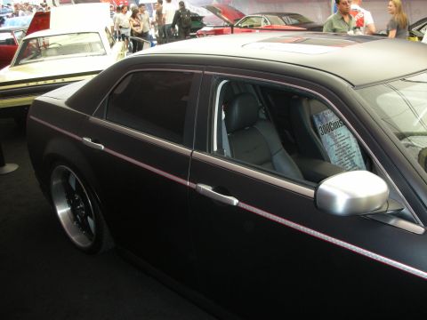 1998 Ford Crown Victoria - Driver-side view of a matte-black Chrysler 300 with black-leather interior, silver mirror and a spec card in the window at an indoor show.