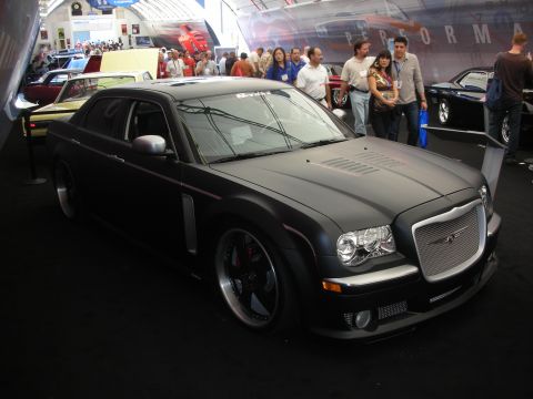 1998 Ford Crown Victoria - Matte-black custom Chrysler 300 sedan on dark wheels with a bright chrome mesh grille, displayed before a crowd at an auto show.