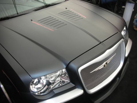 1998 Ford Crown Victoria - Close-up of a matte-black Chrysler 300 nose showing the louvered hood, winged emblem and bright chrome mesh grille.