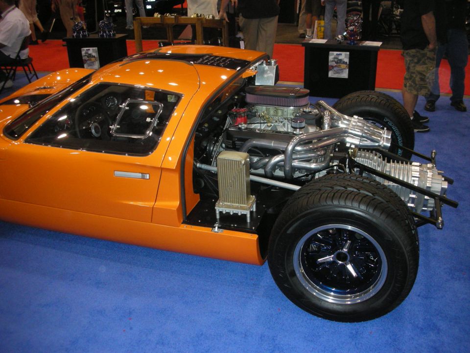 Orange mid-engine sports car cutaway showing V8 and chassis on blue carpet