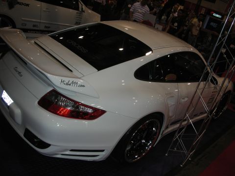 1998 Ford Crown Victoria - White Porsche 911 Turbo coupe, rear three-quarter view with whale-tail spoiler and split chrome wheels at an indoor show.